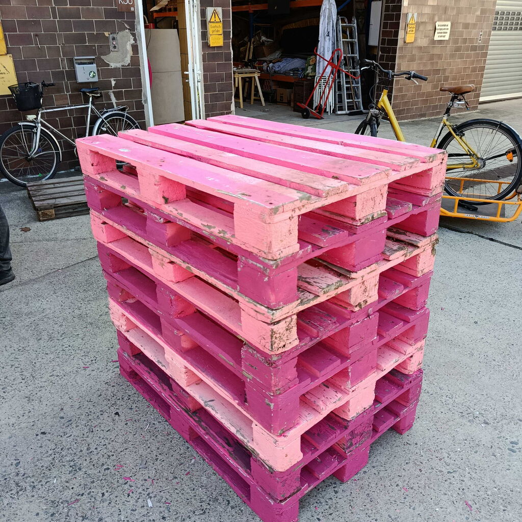 Gestapelte, pink lackierte Holzpaletten auf einem Betonboden vor einer Backsteinwand mit Fahrrädern im Hintergrund